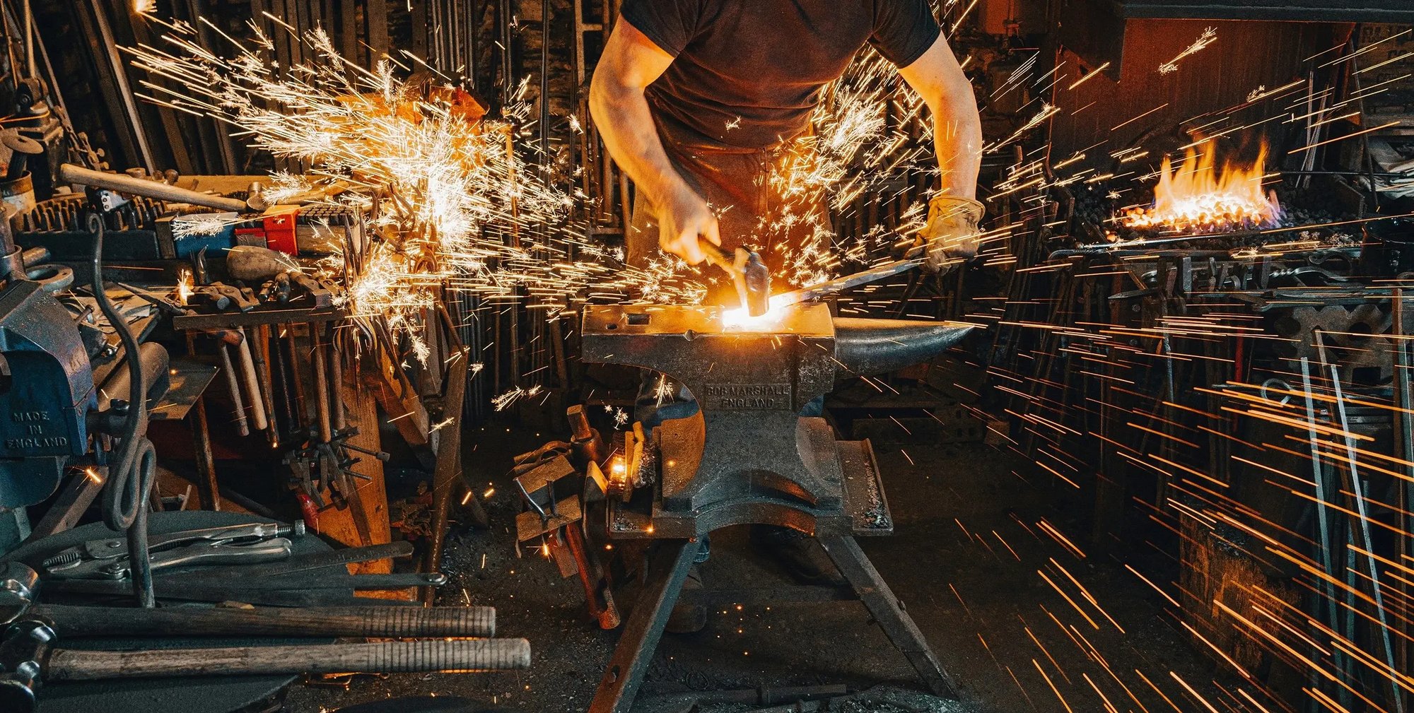 A blacksmith in a foundry hammers a steel rod on an anvil. Sparks shoot out from the impact.