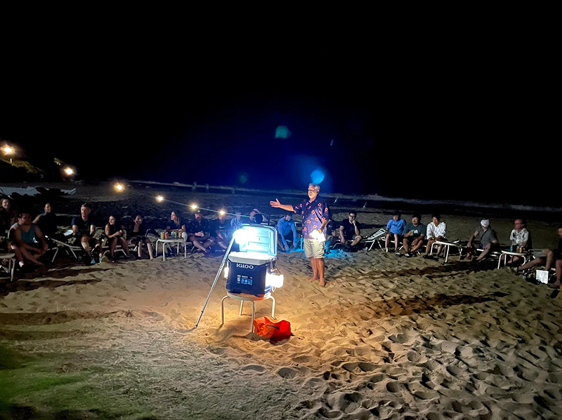 A Digital Foundry employee stands holding a glowing ball and speaking in the middle of a circle of other software consultants and engineers on a beach at night.